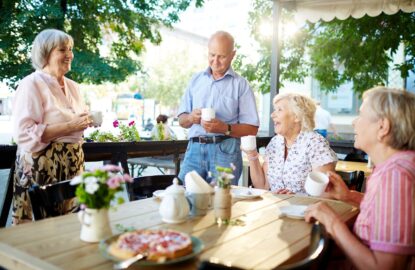 Cheerful senior friends celebrating holiday in outdoor cafe while one of them making toast; teapot and tasty pie standing on wooden table